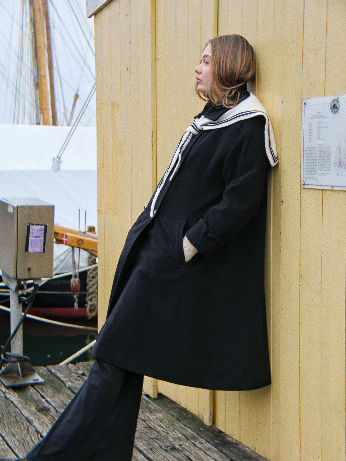 EIn Model lehnt an einer gelben Wand, trägt einen schwarzen Mantel mit Aglaea Studio Schal im maritimen Stil